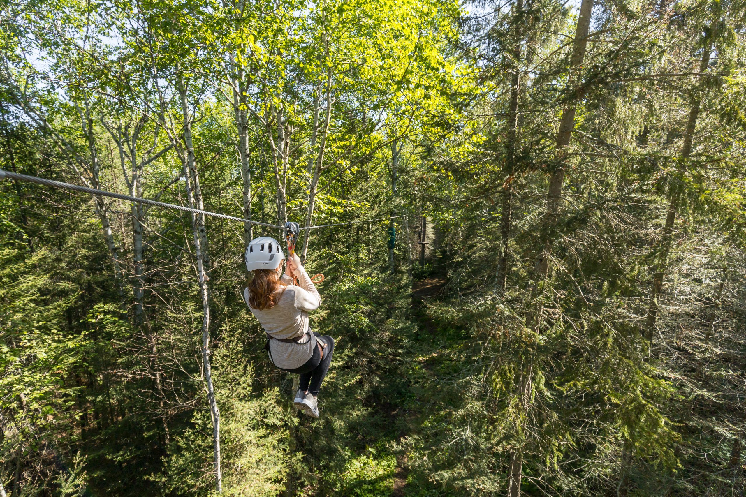 Une femme portant un casque de sécurité est attachée et suspendues dans les airs grâce à une tyrolienne. La femme est entourée par la cime des arbres. Visiter le Parc-Aventure Joannès est une belle activité à faire en famille en Abitibi-Témiscamingue. 